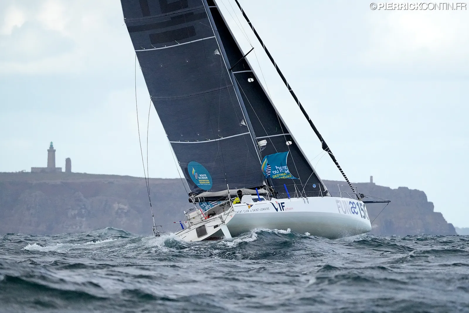 Le Class40 sous voiles noires, devant une côte rocheuse
