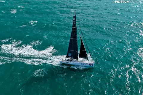 Le Class40 vu du ciel, sous voiles noires, sur une mer turquoise