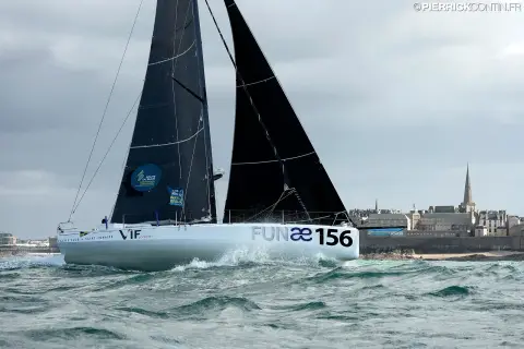 Le Class40 de Loïc Féquet en mer, sous voiles noires, coque blanche portant le nom FUNAE, devant une côte aux bâtiments de pierre