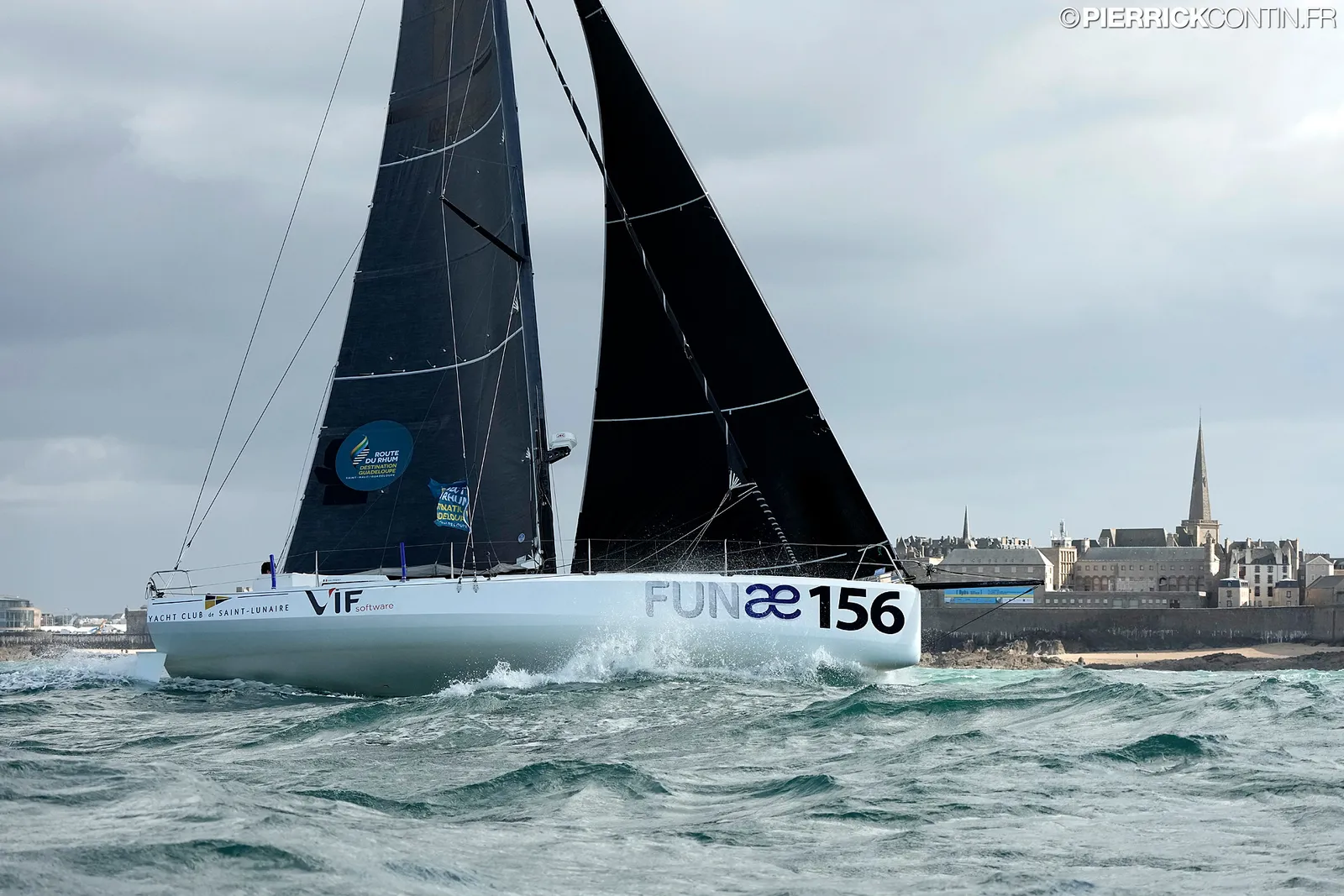 Le Class40 de Loïc Féquet en mer, sous voiles noires, coque blanche portant le nom FUNAE, devant une côte aux bâtiments de pierre